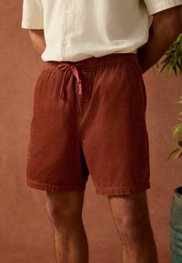 Shorts en velours côtelé couleur rouille avec une taille élastique et un cordon de serrage réglable. Présente un tissu texturé et une coupe droite.