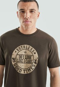 Brązowa bawełniana koszulka z okrągłym wzorem, na którym znajduje się napis "RUTHLESS DO I LOOK LIKE I CARE GOOD TIMES" w postarzanym beżowym kolorze.