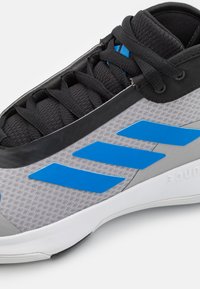 Scarpa sportiva grigia con accenti neri e design a strisce blu. Presenta una tomaia texturizzata, suola in gomma e chiusura con lacci.