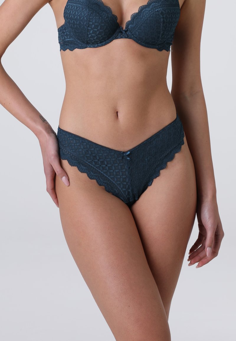 Completo di lingerie in pizzo color teal scuro, caratterizzato da un reggiseno stampato con bordi scallop e un perizoma abbinato con un fiocco decorativo e un design testurizzato.