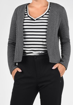 Nainen, jolla on yllään harmaa, teksturoitu bleiseri, mustavalkoraidallinen V-kaula-aukollinen paita ja mustat housut, kädet taskuissa.