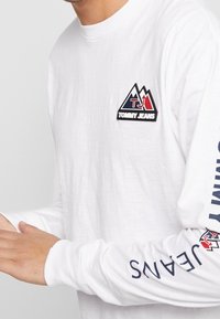 Vit långärmad t-shirt i bomull, med en färgglad bergslogo på bröstet och "JEANS" tryckt i marinblått på ärmen.