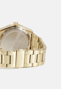 Reloj de acero inoxidable dorado con una correa texturizada, que presenta una caja redonda, una esfera minimalista y un logo grabado en el cierre.