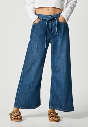 Jeans de mezclilla azul de pierna ancha con un cinturón de tela, combinados con sandalias marrones de hebilla y un top blanco de manga larga y corto. Manos en los bolsillos.