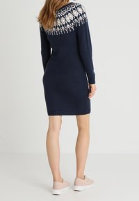 Femme portant une robe tricotée bleu marine avec un encolure en motif blanc et beige, associée à des baskets rose clair, se tenant de dos.