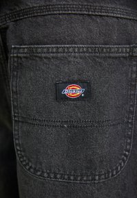 Svart denimt stoff med en baklomme som har et rektangulært Dickies-logo merke i rødt, gult og blått. Synlige sømdetaljer.