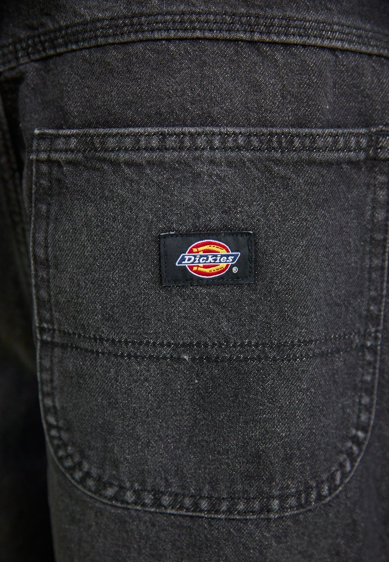 Schwarzer Denim-Stoff mit einer Gesäßtasche, die ein rechteckiges Dickies-Logo-Patch in Rot, Gelb und Blau aufweist. Sichtbare gestickte Details.