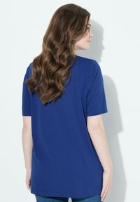 Žena s dlouhými vlnitými hnědými vlasy, oblečená v modrém tričku s krátkým rukávem, viděná zezadu na bílém pozadí.