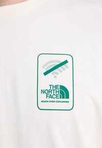 Hvid bomulds t-shirt med et grønt rektangulært logo, der viser "THE NORTH FACE" og et Wi-Fi symbol, der er krydset ud, sammen med sætningen "NEVER STOP EXPLORING."