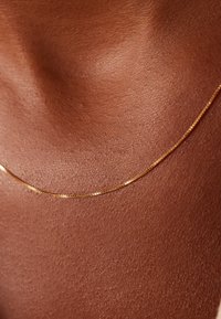 Gouden kettinghanger gepresenteerd tegen een gestructureerde bruine huid, met een verfijnd, dun ontwerp met een gladde oppervlakte en subtiele glans.