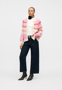 Femme aux cheveux tressés portant un cardigan rose à motifs, un haut blanc, un jean large sombre, une ceinture noire avec une boucle en forme de cœur, et des bottines noires.