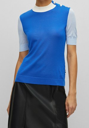 Vrouw draagt een blauw topje met korte mouwen, witte kraag en knopen op de schouder, gecombineerd met een zwarte leren rok.