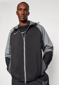 Veste coupe-vent Nike noire et grise avec fermeture éclair, capuche ajustable et tissu léger et texturé. Logo sur la poitrine.