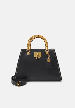 Sac à main trapèze noir avec poignées style bambou, ferrures dorées, coutures visibles, et bandoulière amovible sur fond blanc.