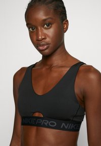 Black sports bra made of smooth fabric, featuring a deep V-shaped neckline, cutout detail, and a wide elastic band with "NIKE PRO" text.