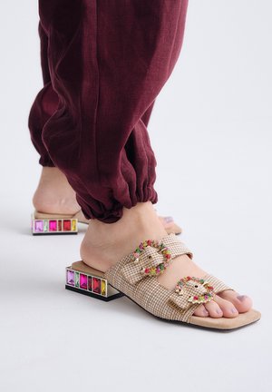 Pies con sandalias beige sin cierre, con hebillas adornadas con joyas y tacones anchos decorados con gemas de colores, combinadas con pantalones burdeos.