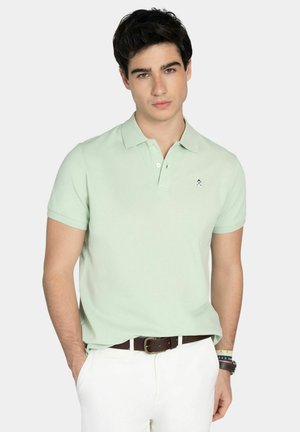 Joven con camisa tipo polo verde claro con un pequeño logo, pantalones blancos, cinturón marrón y pulseras, posando frente a un fondo liso.