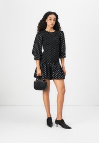 Kvinna stående i en svart prickig klänning med puffärmar, håller en svart handväska och bär svarta snörda kitten heels.