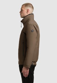 Brown insulated jacket with a high collar, zippered front, side pockets, and a black logo patch on the shoulder. Smooth finish, casual fit.