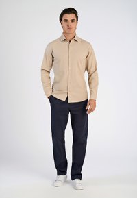 Camicia beige a maniche lunghe con bottoni, abbinata a pantaloni blu navy e sneakers bianche. La camicia presenta un colletto e una texture liscia.
