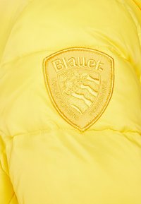 Giacca a vento gialla con finitura texturizzata presenta un logo in rilievo al centro. Il materiale è lucido, morbido e leggero.