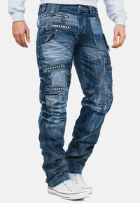 Pantaloni cargo in denim di colore blu scuro con molteplici tasche, cuciture a contrasto e dettagli decorativi in metallo. Abbinali a sneakers bianche.