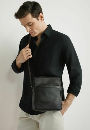 Mężczyzna w czarnej koszuli i beżowych spodniach, trzymający pasek czarnej skórzanej torby na ramię na jednolitym jasnym tle.