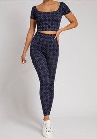 Navyblauw cropped top met korte mouwen, gecombineerd met high-waisted leggings, beide met een herhalend logo patroon, gestyled met witte sneakers.