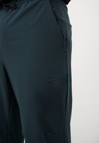 Tumši gremuanā Nike sporta bikses ar gludu tekstūru, elastīgu jostasvietu, šūtām sānu kabatām un toņos atbilstošu logo uz kreisā kājas.