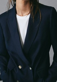 Donna che indossa un blazer doppiopetto blu navy con bottoni metallici sopra una maglietta bianca a girocollo, mani nelle tasche, tagliato a livello del collo e dei fianchi.