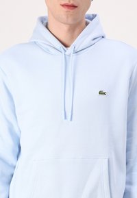 Sudadera azul clara hecha de una tela suave, con un bolsillo tipo canguro y un logo de cocodrilo bordado en el lado izquierdo del pecho.
