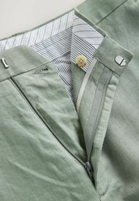 Grüne Leinen-Shorts mit Reißverschluss, Knopfverschluss und gestreiftem Innenfutter. Sie verfügen über einen moderaten Glanz und einen strukturierten Stoff.