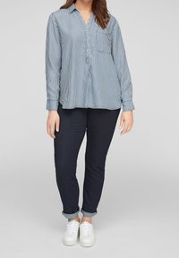Camicia a righe blu e bianche con colletto, abbinata a jeans di jeans scuri e sneakers bianche. La camicia ha una sola tasca sul petto.