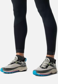 Grau und schwarze Trailrunning-Schuhe mit blauen und rosa Akzenten, strukturierten Sohlen und reflektierenden Details. Kombiniert mit schwarzen Leggings.