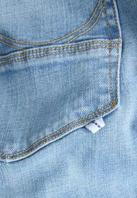 Tessuto di denim azzurro chiaro con texture visibile, cuciture in oro e un piccolo etichetta bianca sulla cucitura della tasca.