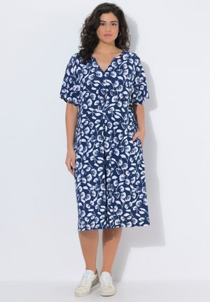 Kvinde stående iført en knælængde, mørkeblå kjole med hvidt blomstermønster, korte ærmer, V-hals og hvide sneakers, hænderne i lommerne.