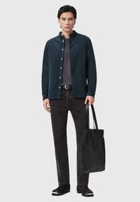Camicia button-up in corduroy verde scuro sopra una t-shirt grigia, abbinata a jeans neri e una borsa a tracolla nera con texture. Calzature lisce con un accento in argento.
