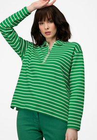 Maglia a maniche lunghe a righe verdi con colletto zip. Presenta righe orizzontali color crema e vestibilità rilassata, abbinata a pantaloni verdi.