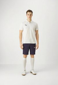 Valge polomehe särk musta logoga, tumelilla lühikesed püksid, valged põlveni ulatuvad sokid ja valged tossud beežide talladega. Klassikaline spordiriietus.