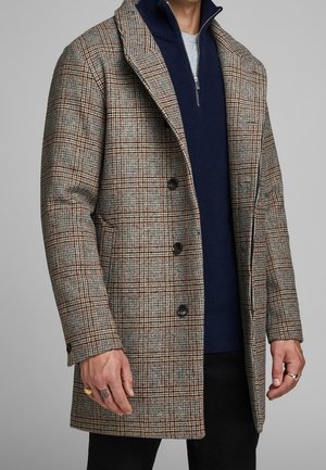 Braun karierter Mantel aus strukturierter Stoff mit einem Reverskragen, Knopfverschluss und einem kontrastierenden navyblauen Reißverschlusspullover darunter.