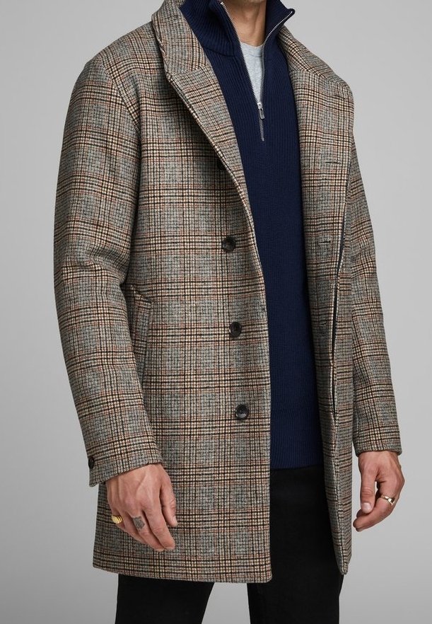 Cappotto a quadri marroni con tessuto strutturato, dotato di colletto a revers, chiusura a bottoni e un maglione blu navy a zip in contrasto sotto.