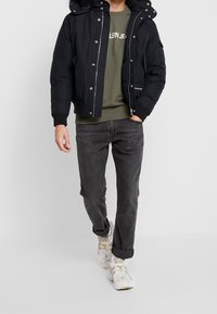 Giubbotto imbottito nero con cappuccio foderato in pelliccia, abbinato a una felpa verde e jeans grigi. Presenta chiusure a pressione e con cerniera, insieme a scarpe da ginnastica sportive.