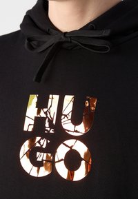 Sudadera negra con un tejido texturizado. Presenta un gran logo reflejante "Hugo" en dorado con un patrón agrietado. Detalle de capucha con cordones visible.