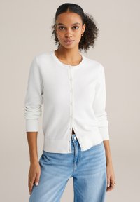 Cardigan alb tricotat, cu guler rotund, nasturi frontal și manșete cu reiat. Purtați peste blugi albastru deschis, având un model texturat.