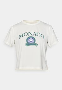 Fehér pamut póló zöld "MONACO" felirattal, kerek logó kék díszítésekkel, rövid ujjakkal és oldalhasítékos cropped fazonnal.