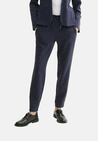 Joggers ajustés bleu marine avec une taille à cordon, deux poches latérales et une texture lisse, associés à des chaussures noires à lacets.