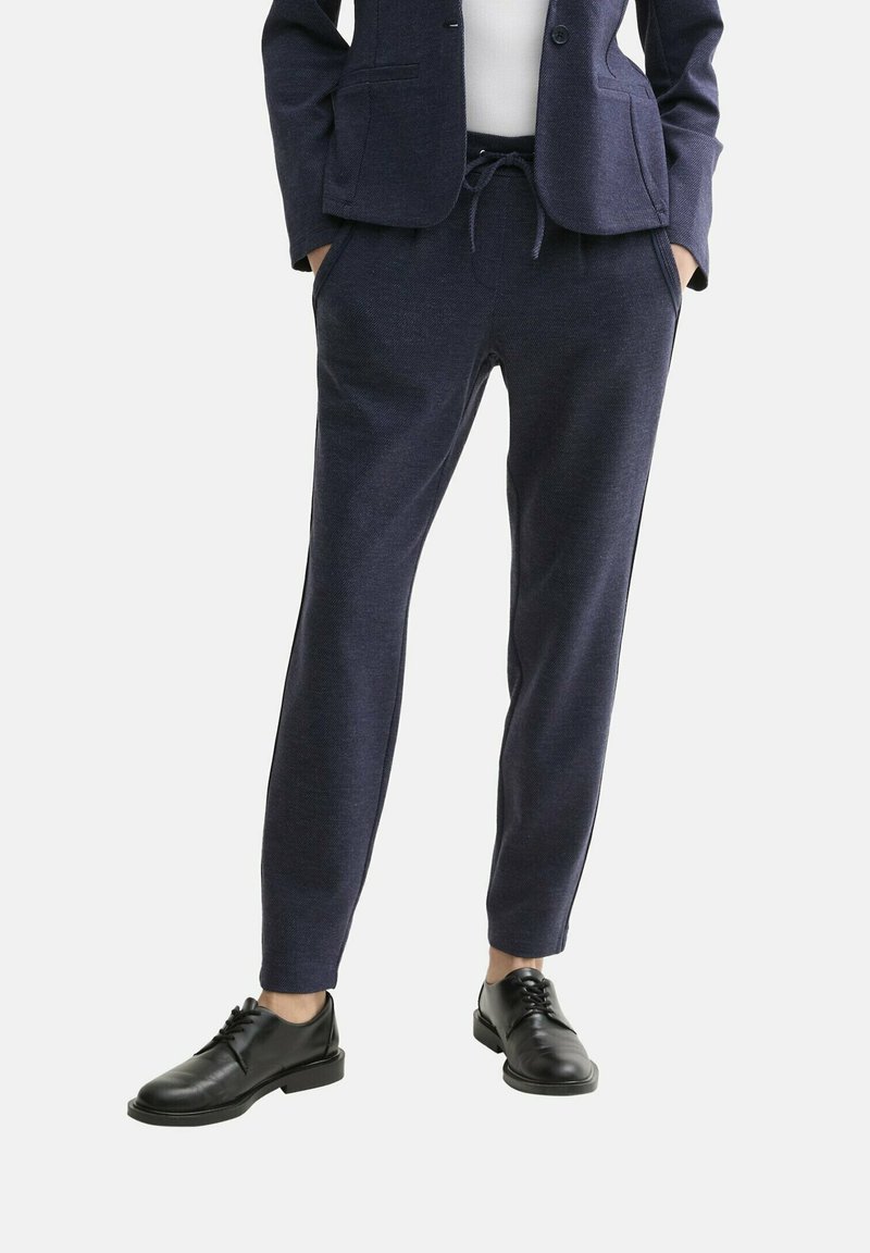 Joggers ajustés bleu marine avec une taille à cordon, deux poches latérales et une texture lisse, associés à des chaussures noires à lacets.