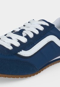 Gros plan d'une chaussure de sport bleue avec des lacets blancs, une bande latérale blanche et une semelle marron texturée sur fond blanc.