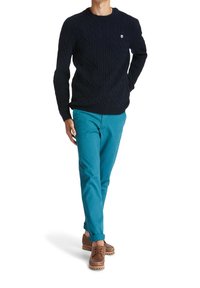 Pull en maille torsadée bleu marine associé à un chino vert sarcelle et des chaussures marron à lacets. Le pull présente un col rond et un détail de logo discret.