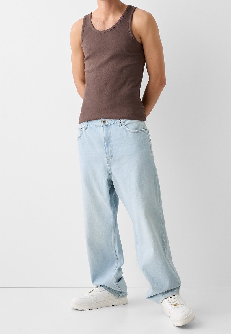 Homme portant un débardeur marron, un jean large bleu clair et des baskets blanches, se tenant contre un fond uni.